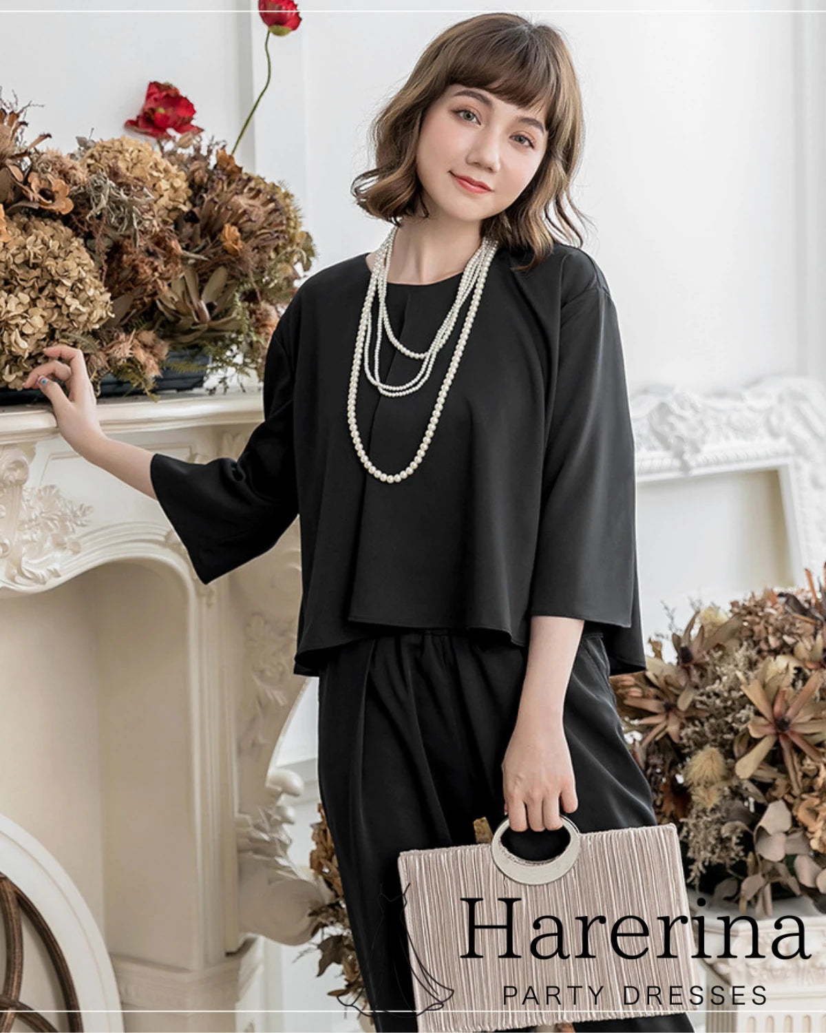黒のケープ風トップスとパンツのセットアップを着た女性が、パールのネックレスとフリンジクラッチバッグを持って立っているコーディネート写真
