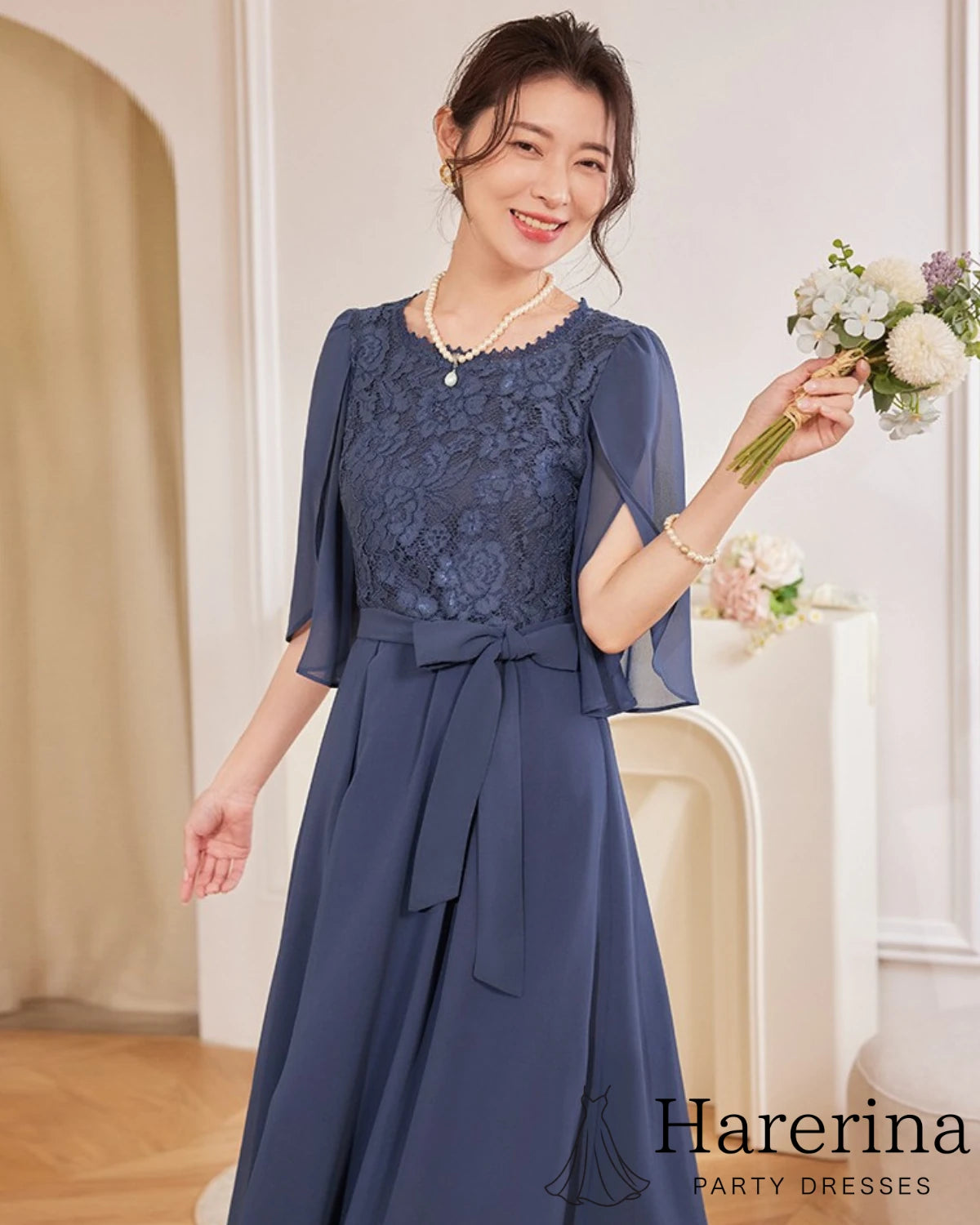 ダークブルーの軽やかなシフォン×ウエストリボンのエレガントドレスを着た女性が小さな花束を手に微笑んでいる様子
