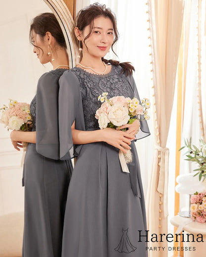 ダークグレーの軽やかなシフォン×ウエストリボンのエレガントドレスを着た女性が、花束を両手で持って鏡の前で微笑んでいる様子
