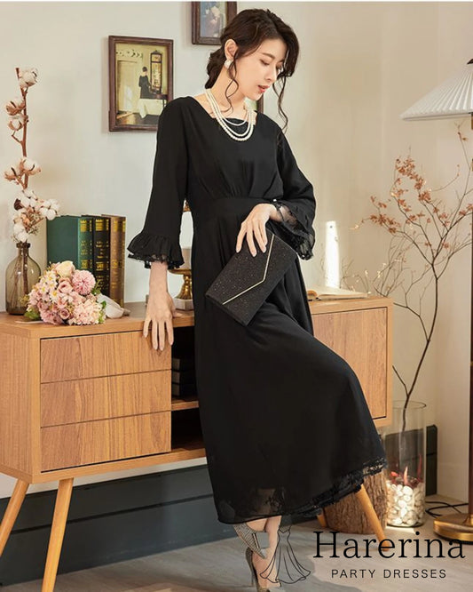 黒のレース袖×フレアロングワンピースを着た女性がキャビネットに寄りかかり、クラッチバッグを持って上品に佇むフォーマルコーデ写真。
