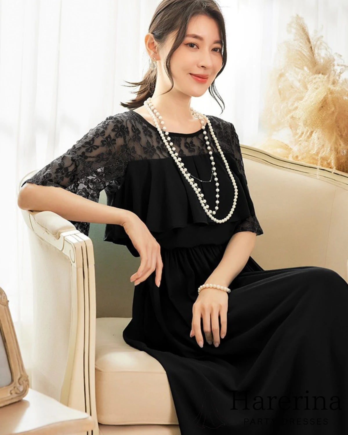 ブラックのフェミニンレーストップ×エアリーシフォンロングワンピースを着た女性が、ソファに座って微笑む様子を写した画像
