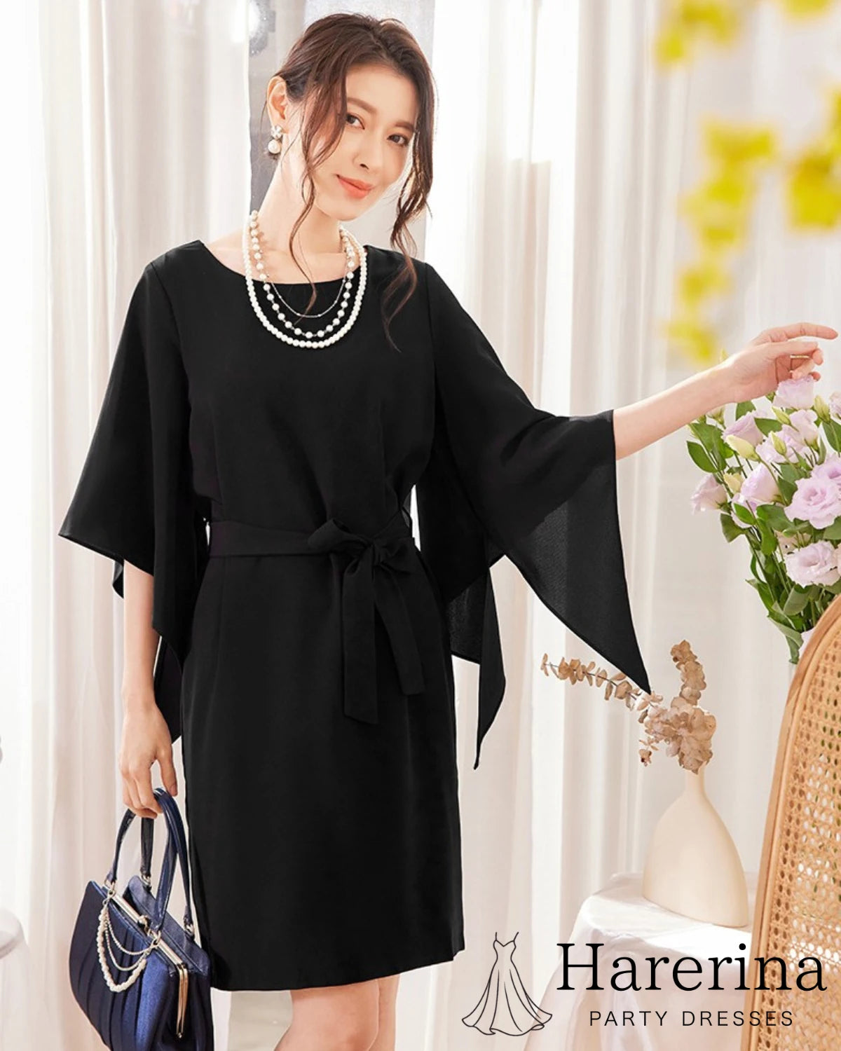 ブラックのフレアスリーブ×ウエストリボン風デザインワンピースを着た女性が花に手を添えて優しく微笑む全身写真。
