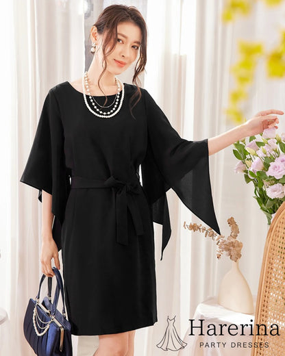 ブラックのフレアスリーブ×ウエストリボン風デザインワンピースを着た女性が花に手を添えて優しく微笑む全身写真。
