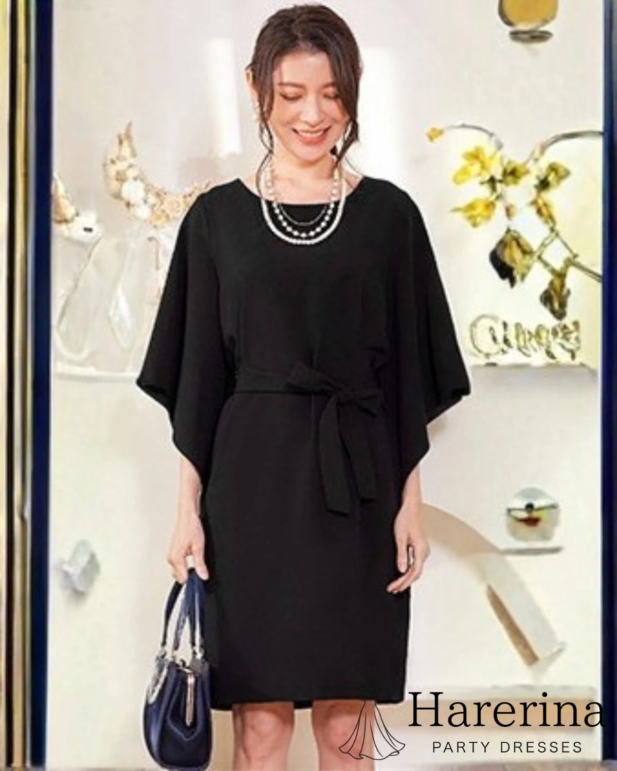 ブラックのフレアスリーブ×ウエストリボン風デザインワンピースを着た女性がネイビーのバッグを手に微笑んで立つ全身写真。
