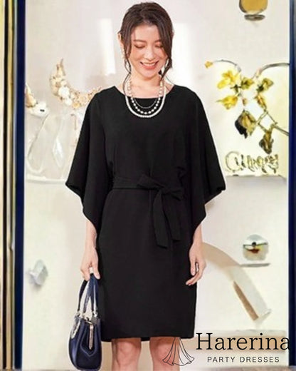 ブラックのフレアスリーブ×ウエストリボン風デザインワンピースを着た女性がネイビーのバッグを手に微笑んで立つ全身写真。
