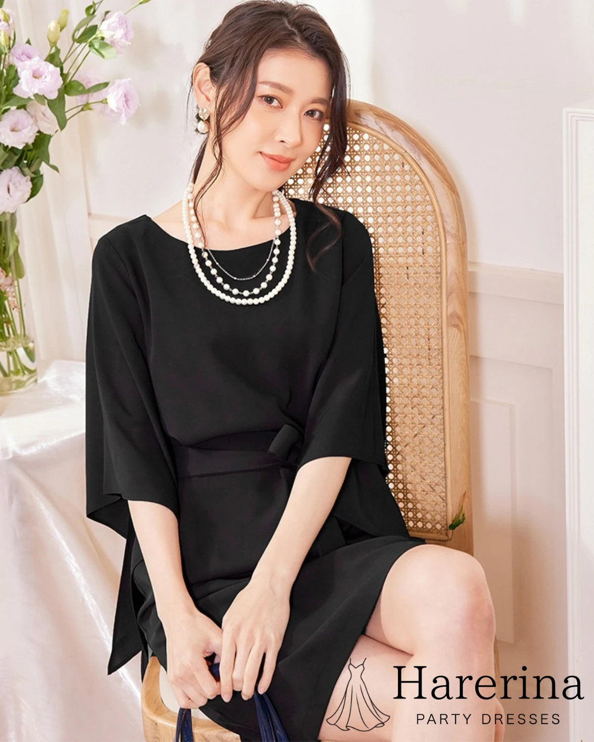 ブラックのフレアスリーブ×ウエストリボン風デザインワンピースを着た女性が椅子に座って上品に微笑む写真。
