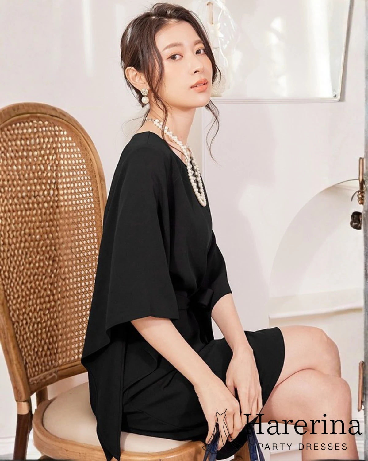 ブラックのフレアスリーブ×ウエストリボン風デザインワンピースを着た女性が椅子に座り横向きで視線を向けている写真。
