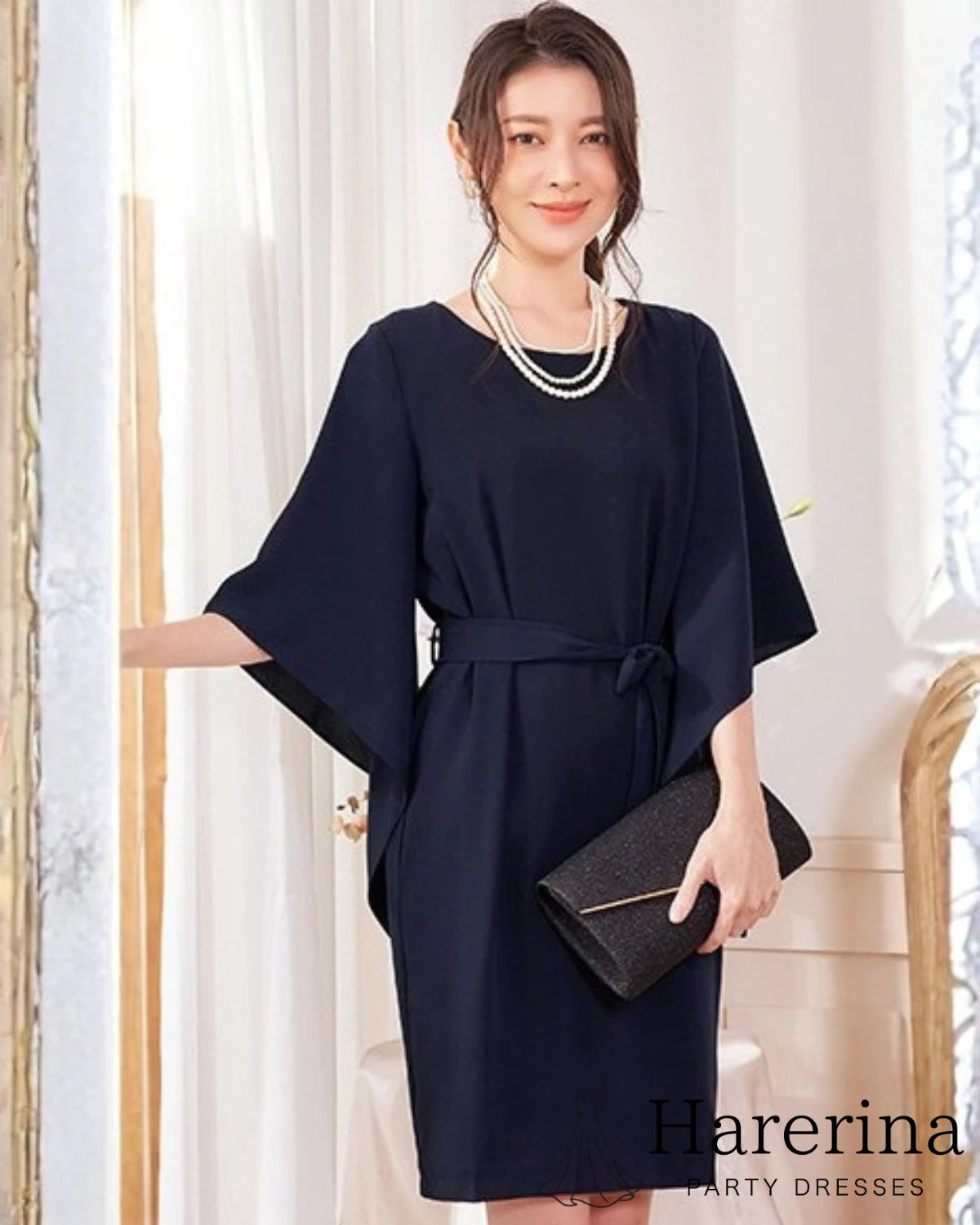 ネイビーのフレアスリーブ×ウエストリボン風デザインワンピースを着た女性が正面に向かって微笑みながらクラッチバッグを持つ全身写真。
