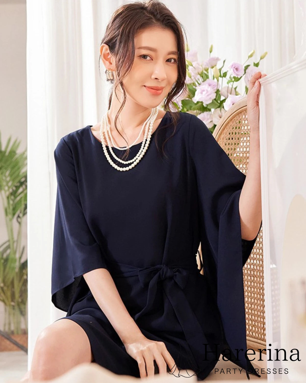 ネイビーのフレアスリーブ×ウエストリボン風デザインワンピースを着た女性が椅子に座りながら微笑む上品な雰囲気の写真。
