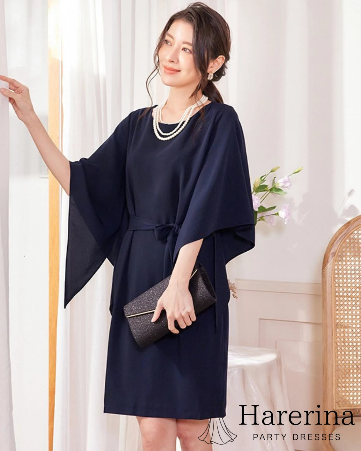 ネイビーのフレアスリーブ×ウエストリボン風デザインワンピースを着た女性がカーテンに手を添えて横向きに立つ上品な雰囲気の写真。
