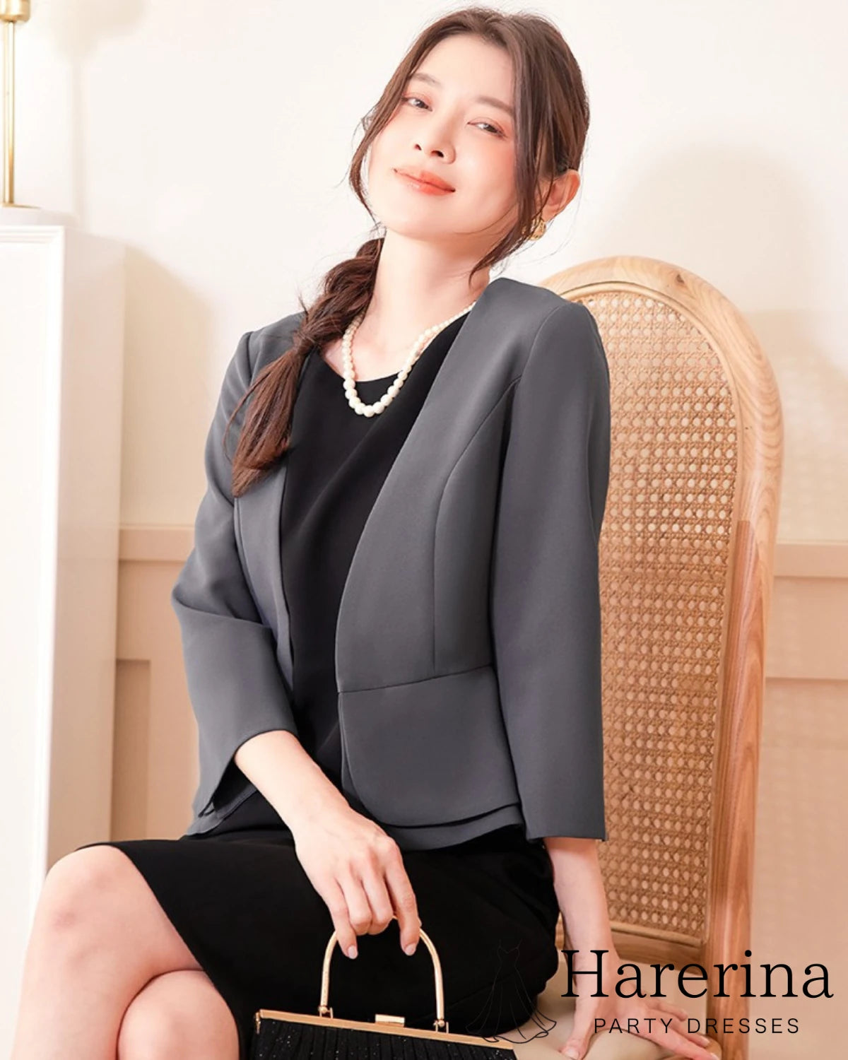 ダークグレーのペプラム風フォーマルジャケットを着用した女性が椅子に座って微笑む上半身寄りの写真
