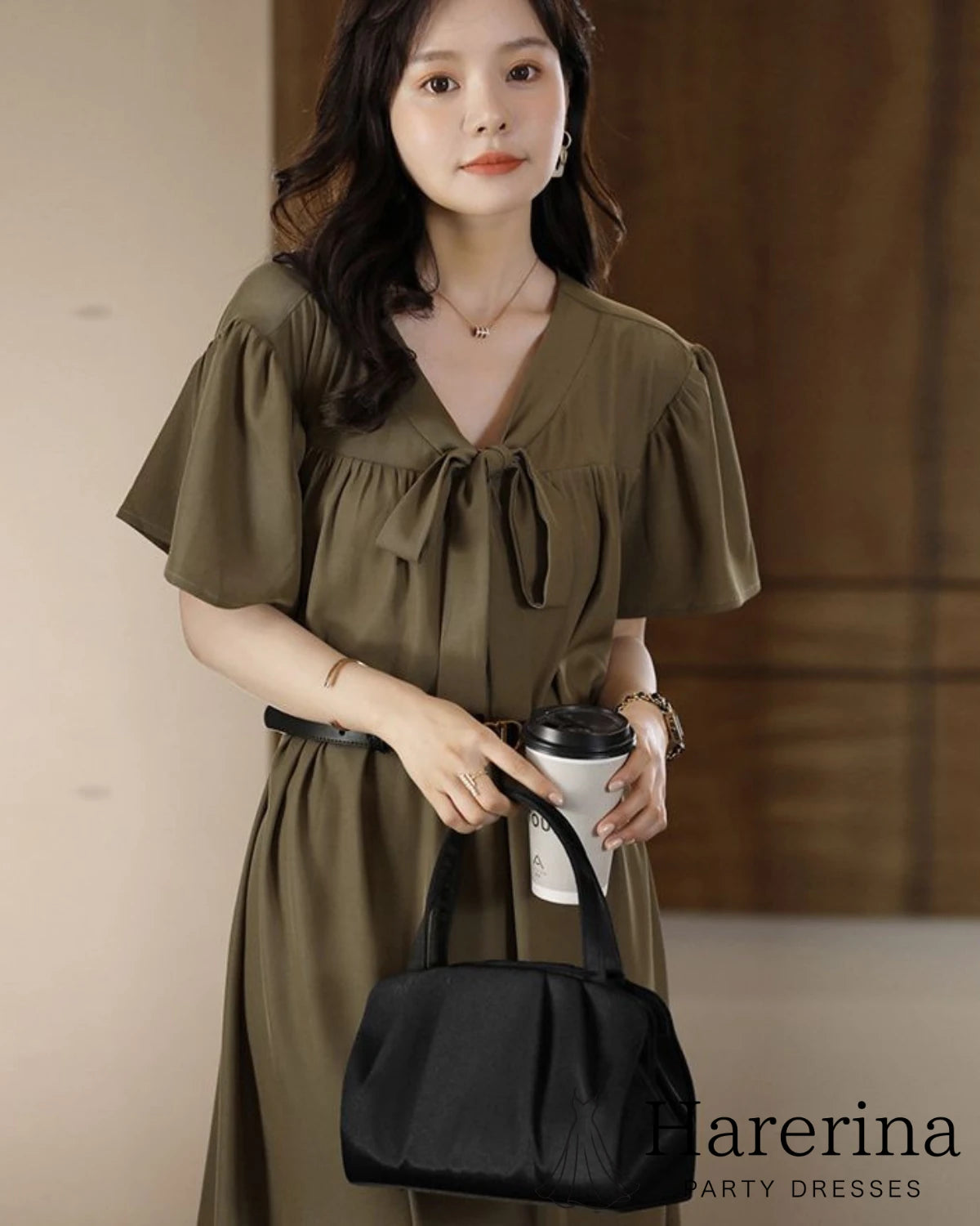 カーキ色のワンピースを着た女性がコーヒーカップを持ち、ブラックのプリーツデザインパーティーバッグを腕に掛けている上半身寄りの着用イメージ
