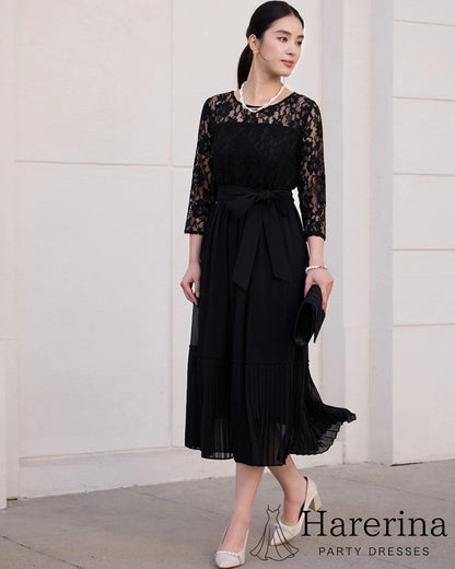 ブラックのレースシフォンドレスを着た女性が黒いクラッチを持って屋外で立つ全身正面イメージ
