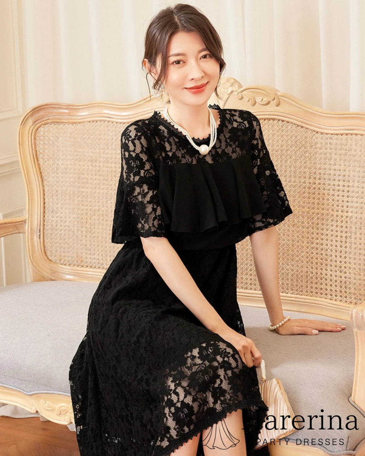 フリルレースが華やかなアシンメトリードレス（ブラック）を着た女性がソファに座り上品に微笑む姿

