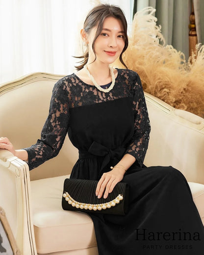 ブラックのレーススリーブ×ウエストリボン ロングワンピースを着た女性が、ソファに座りクラッチバッグを持って上品に微笑む商品画像。
