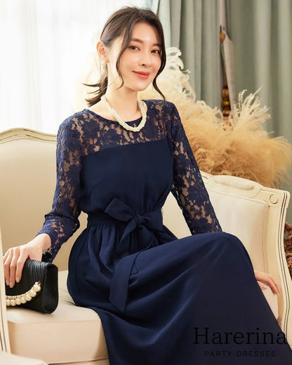 ネイビーのレーススリーブ×ウエストリボン ロングワンピースを着た女性が、ソファに座り上品に微笑む商品画像。

