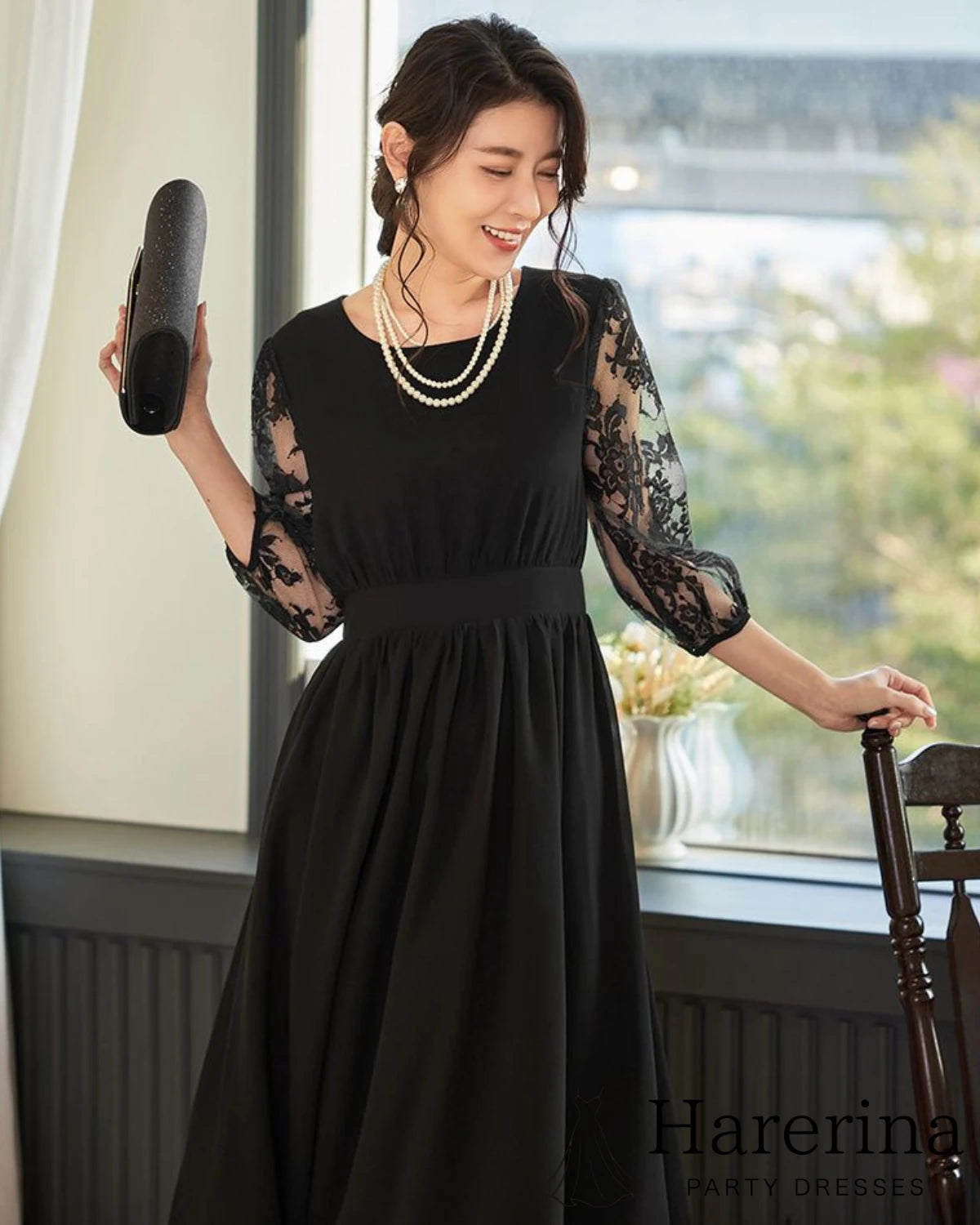 ブラックのレース袖プリーツワンピースを着た女性がクラッチバッグを持って笑顔を見せている上半身カット
