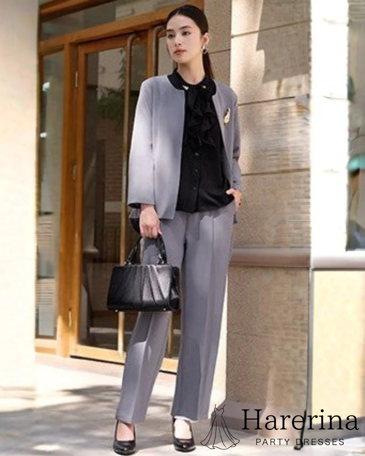 グレーのノーカラージャケット×パンツセットアップを着た女性モデルが黒バッグを持って立つ全身コーデ。黒ブラウスを合わせた落ち着いたフォーマルスタイル。
