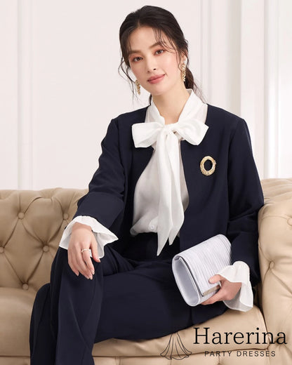 ネイビーのノーカラージャケット×パンツセットアップを着た女性モデルがソファに座る上半身コーデ。白のボウタイブラウスとゴールドブローチを合わせた上品なスタイル。
