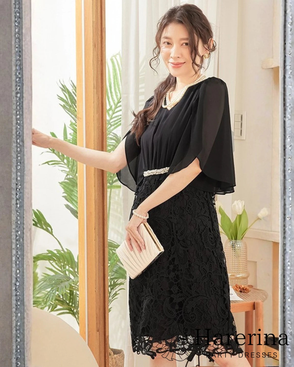 ブラックカラーのパールウエストマーク×膝丈レースドレスを着用した女性を正面から写した画像。シフォン袖とレーススカートが上品で、クラッチバッグを持ったエレガントなスタイル

